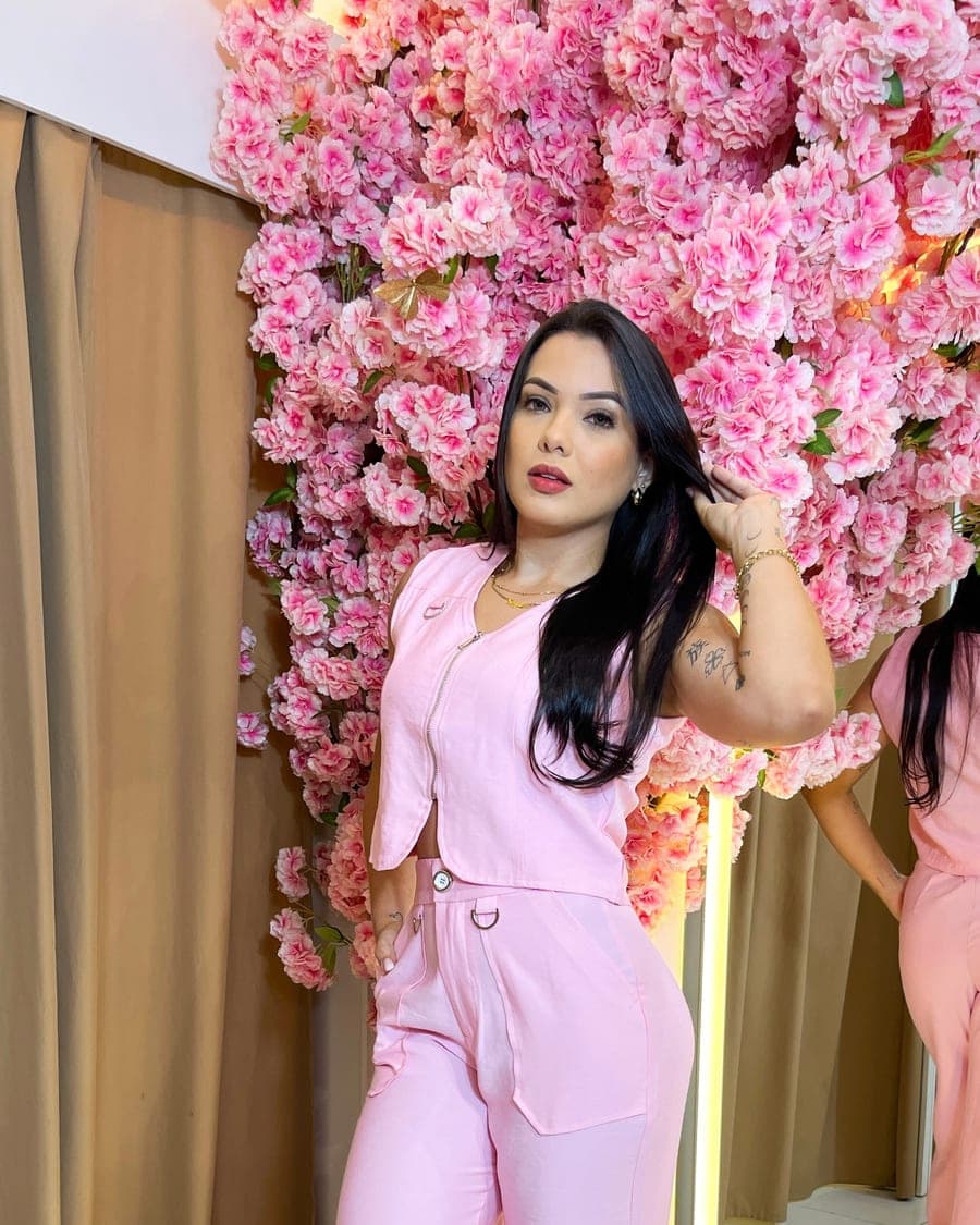Sayuri Castro com conjunto rosa de colete e calça, diante de uma parede de flores