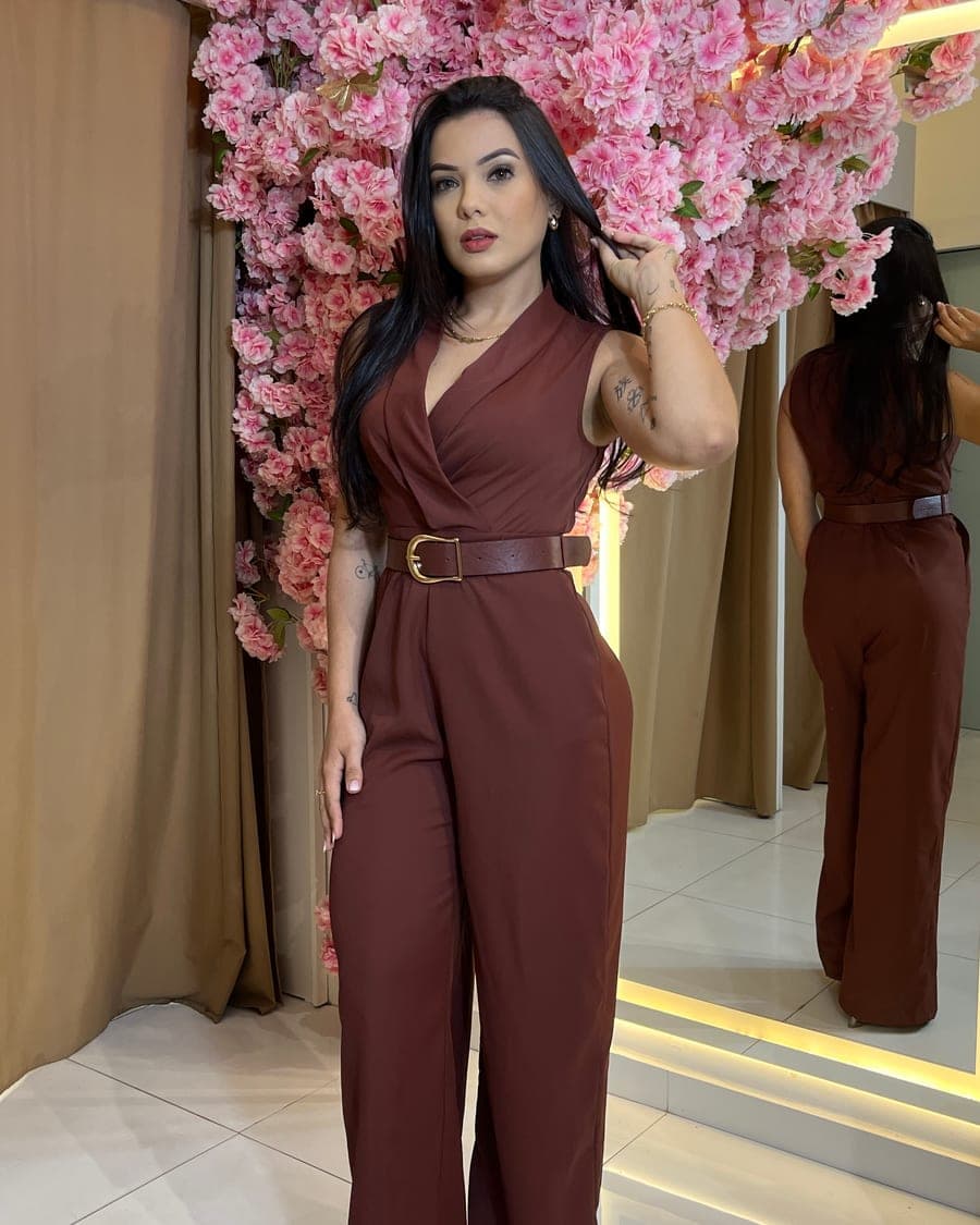 Sayuri Castro com macacão marrom de cinto, diante de uma parede de flores